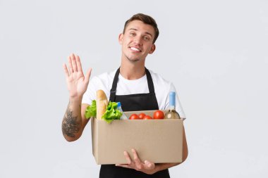 Perakende, market alışverişi ve teslimat konsepti. Dost canlısı yakışıklı satıcı sebzelerle dolu bir kutu yaptı, yiyecek siparişi verdi, merhaba demek için el salladı ve sebzeli bir kutu tuttu, beyaz arka plan.