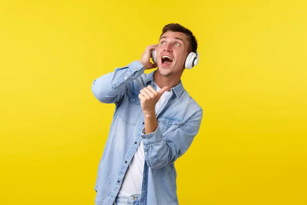 Stile di vita, vacanze estive, concetto tecnologico. Bello spensierato dj maschile, ragazzo che gode di un suono impressionante di musica, mostrando pollici in alto a sinistra, indossando cuffie wireless, sfondo giallo — Foto Stock