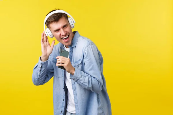 Freizeit-, Technologie- und Anwendungswerbekonzept. Schöner junger kaukasischer Mann hat Spaß, spielt Karaoke-App auf Handy, benutzt Smartphone als Mikrofon und singt mit Kopfhörer — Stockfoto