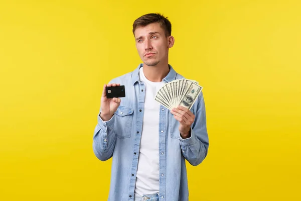 Pensativo hombre guapo rubio serio que muestra la tarjeta de crédito y el dinero, mirando hacia otro lado pensando cómo invertir dinero, sopesando lo que comprar durante las compras, de pie fondo amarillo — Foto de Stock
