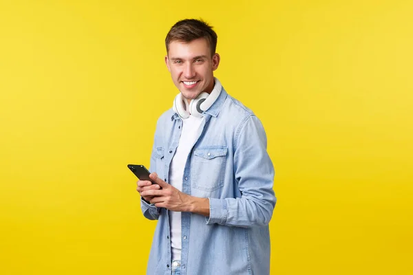 Boş zaman, teknoloji ve uygulama reklam kavramı. Yakışıklı adam cep telefonu ve kulaklık kullanırken gülümsüyor, mesajlaşıyor, araba paylaşımı uygulaması kullanıyor, mesajlaşıyor, sarı arka plan — Stok fotoğraf