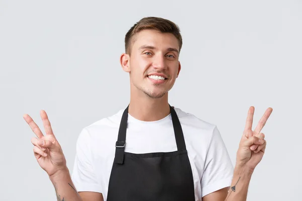 Kawiarnia i restauracje, właściciele kawiarni i koncepcja sprzedaży detalicznej. Close-up przyjazny wesoły barista, kelner w czarnym fartuchu, pokazując znak pokoju i uśmiechając się szeroko, powitanie gości, białe tło — Zdjęcie stockowe