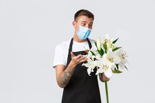 Coronavirus, social distancing business covid-19 pandemic concept. Friendly charismatic florist passionatly describe bouquet, selling flowers, holding white lilies, wear medical mask