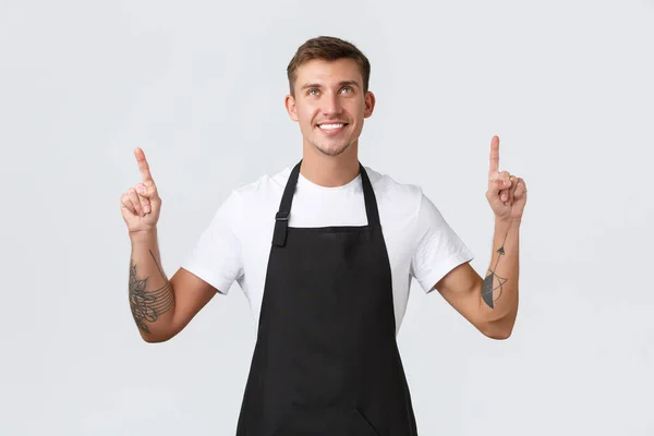 Small business owners, coffee shop and staff concept. Handsome barista, cafe businessman working at counter in black apron, looking and pointing fingers up with pleased smile, white background — Stock Photo, Image
