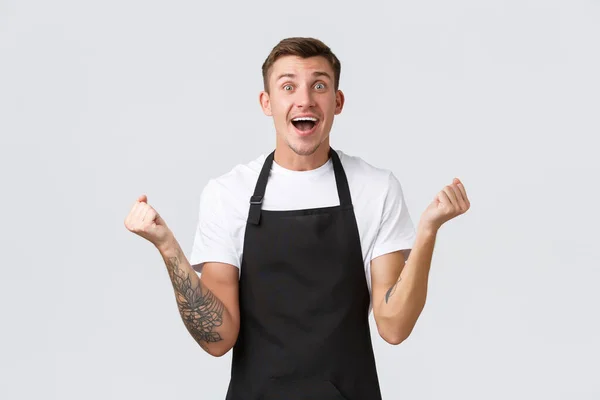 Anställda, livsmedelsbutiker och kafékoncept. Glad och lättad leende stilig barista, servitör i svart förkläde, knytnäve pump i glädje, triumferande seger eller stora positiva nyheter — Stockfoto