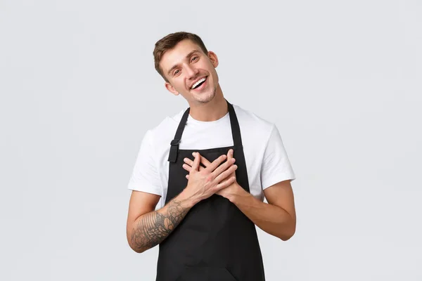 Small business owners, coffee shop and staff concept. Friendly happy and delighted handsome barista, waiter in black apron appreciate cute gesture, tilt head and hold hands on heart pleased — Stock Photo, Image