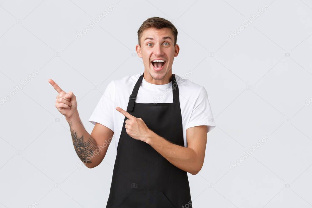 Small business owners, coffee shop and staff concept. Excited happy handsome blond guy, barista in cafe wearing black apron, pointing fingers upper left corner, showing promo discount banner