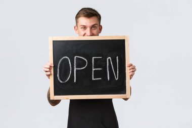 Küçük perakende şirketleri, kafe ve restoran çalışanları konsepti. Yakışıklı, neşeli barista, siyah önlüklü garson açık tabela olduğumuzu gösteriyor, müşterileri bilgilendiriyor, beyaz arka planda duruyor.