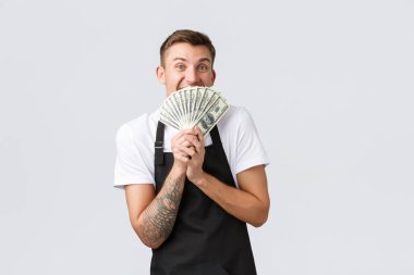 Küçük perakende işi, ödemeler ve çalışanlar konsepti. Kafede ya da restoranda çalışan neşeli, heyecanlı sarışın adam, büyük para gösteriyor, elinde nakit para tutuyor ve neşeyle gülümsüyor, ilk maaşını alıyor.