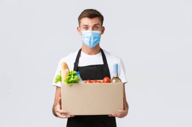 Covid-19, temassız alışveriş ve market teslimatı konsepti. Yakışıklı arkadaş canlısı bir satıcı, tıbbi maskeli bir kurye, marketten alışveriş kutusunu elinde tutuyor, sipariş satıyor, beyaz bir geçmişi var.