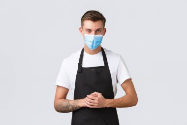 Coronavirus, kafelerde ve restoranlarda sosyal mesafeler, pandemik konsept sırasında iş. Yakışıklı barista, küçük perakende mağazasının satış elemanı siyah önlüklü ve sağlık maskeli kafe misafirleri.