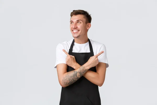 Small business owners, coffee shop and staff concept. Excited handsome smiling barista, waiter in cafe pointing fingers sideways and looking left happy, making choice, have variants, white background — Stock Photo, Image