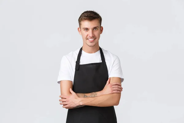 Small business owners, coffee shop and staff concept. Handsome smiling barista in black apron smiling, serving guests in restaurant, prepare order for guests, standing white background — Stock Photo, Image