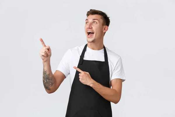 Small business owners, coffee shop and staff concept. Happy surprised smiling handsome guy working in cafe, barista pointing fingers upper left corner, waiter rejoicing over good news — Stock Photo, Image