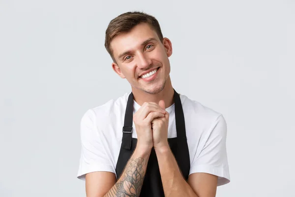 Cafe and restaurants, coffee shop owners and retail concept. Close-up of lovely handsome barista, waiter in black apron, tilt head and hold hands together, smiling with affection and tenderness