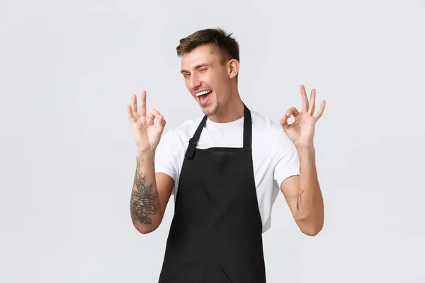 Employees, grocery stores and coffee shop concept. Outgoing cheerful barista, waiter in black apron guarantee quality, showing okay gesture and wink cheeky, approve and recommend cafe — Stock Photo, Image