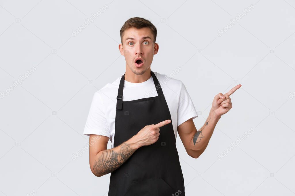 Small business owners, coffee shop and staff concept. Impressed and excited handsome employee in cafe, barista in black apron pointing fingers right and drop jaw shocked, white background