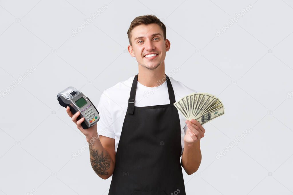 Small retail business, payments and employees concept. Friendly handsome salesman, barista or waiter in black apron giving client POS terminal as customer paying, holding money