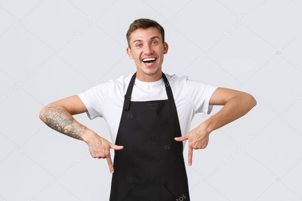 Small business owners, coffee shop and staff concept. Happy friendly barista, waiter in black apron pointing fingers down, guy working in cafe and showing promo or special announcement