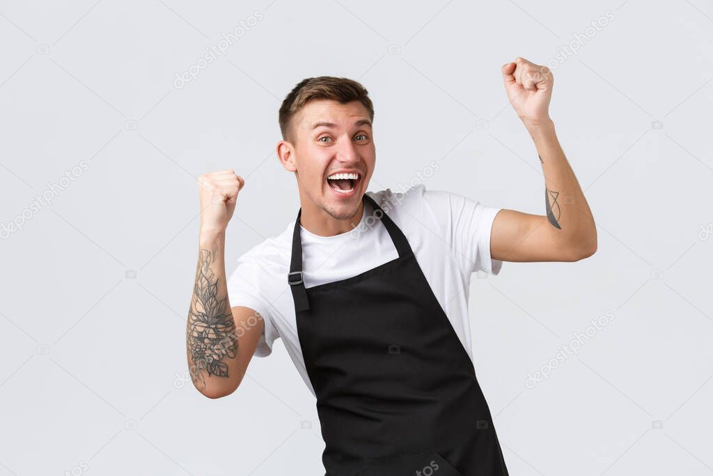 Employees, grocery stores and coffee shop concept. Cheerful grocery store worker, barista in black apron rejoicing over wonderful news, saying yes, winning, raising hands up in triumph