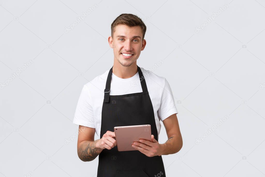Small business, coffee shop and cafe employees concept. Handsome charismatic smiling barista, waiter in black apron, taking customer order in digital tablet, white background