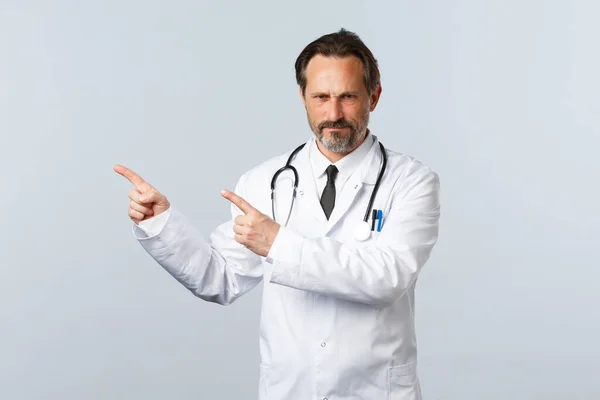 Covid-19, coronavirus outbreak, healthcare workers and pandemic concept. Mad disappointed male doctor in white coat, frowning and pointing fingers left disapproval, condemn smth — Stock Photo, Image