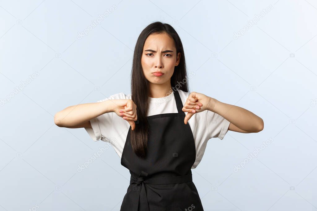 Coffee shop, small business and startup concept. Disappointed gloomy female cafe staff in black apron vote against, show negative judgement. Frowning barista thumbs-down and condemn something