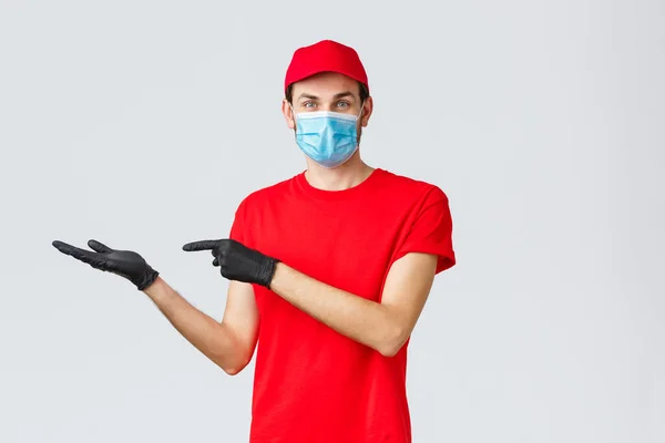 Livraison d'épicerie et de colis, concept covid-19, quarantaine et shopping. Courrier amical en uniforme rouge, masque facial et gants, introduire la promo sur la bannière, pointant à gauche, montrant la publicité — Photo