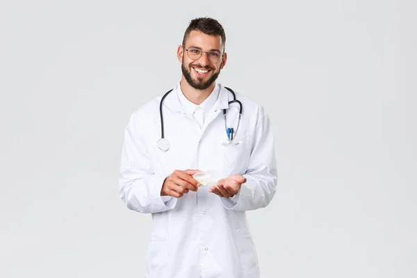 Egészségügyi dolgozók, egészségbiztosítás, világjárvány és kovid-19 koncepció. Jóképű mosolygós orvos fehér köpenyben és szemüvegben, kezében tartja a gyógyszert, tanácsot használ gyógyszert vagy antibiotikumot — Stock Fotó