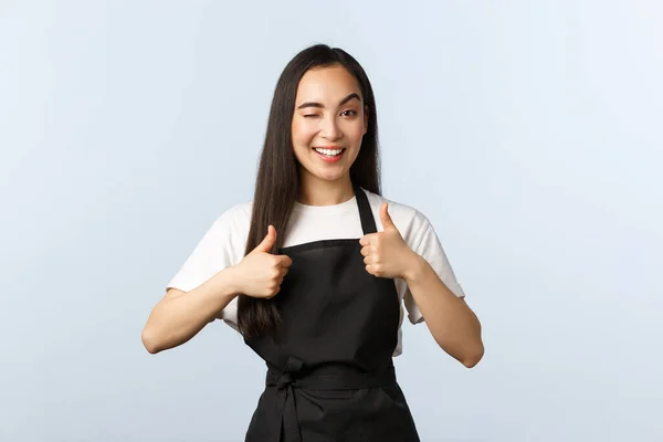 Cafetería, pequeña empresa y concepto de startup. Amistosa chica asiática descarada en delantal negro trabajando como barista en la cafetería o restaurante mostrando el pulgar hacia arriba, guiño y sonriendo, invitando a probar nuevo menú — Foto de Stock