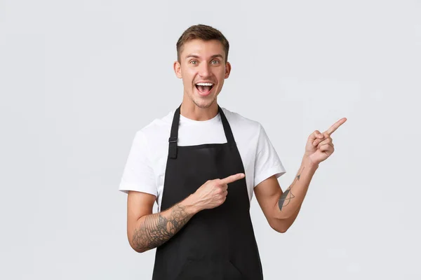 Propietarios de pequeñas empresas, cafetería y concepto de personal. Feliz trabajador a tiempo parcial guapo en la cafetería, camarero o camarero en delantal negro señalando los dedos esquina superior derecha y sonriendo optimista — Foto de Stock