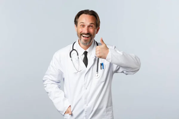 Covid-19, ξέσπασμα του ιού της στέψης, επαγγελματίες υγείας και πανδημία. Ευτυχισμένος χαμογελαστός άνδρας γιατρός με λευκό παλτό χαμογελώντας ευχαριστημένος, δείχνουν αντίχειρες-up, να συστήσει και να προωθήσει τις υπηρεσίες κλινική — Φωτογραφία Αρχείου