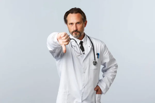 โควิด-19 การระบาดของไวรัสโคโรนา พนักงานสาธารณสุข และแนวคิดการระบาด หมอชายที่ผิดหวังในเสื้อโค้ทสีขาว ไม่พอใจและแสดงนิ้วโป้งลง ไม่เห็นด้วยกับการกระทํา — ภาพถ่ายสต็อก