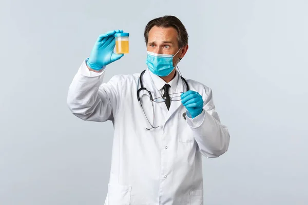 Covid-19: 바이러스 예방, 의료 종사자 및 예방 개념. 마스크와 장갑을 낀 진지 해 보이는 의사가 소변 샘플을 검사하고 사려 깊고 집중적으로 보고 있습니다 — 스톡 사진