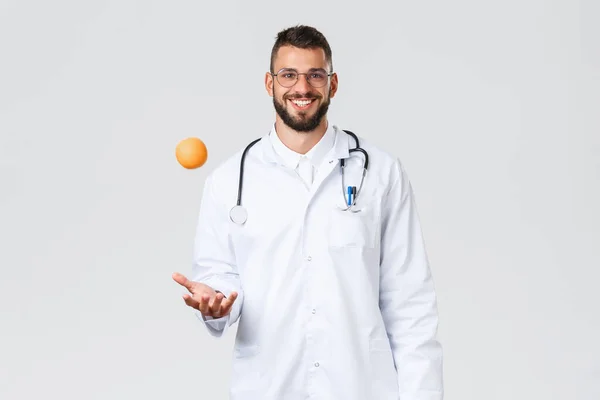 Pracownicy służby zdrowia, ubezpieczenie zdrowotne, laboratorium kliniczne i koncepcja covid-19. Wesoły uśmiechnięty lekarz, lekarz w białym płaszczu, rzut pomarańczowy, polecam jeść zdrowe owoce witamin — Zdjęcie stockowe