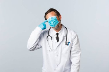 Covid-19, virüsü önleme, sağlık çalışanları ve aşı konsepti. Yorgun ve sıkıntılı doktor Coronavirus 'la savaşıyor, yorgun iş, maske takıyor ve kafasına silah dayıyor.