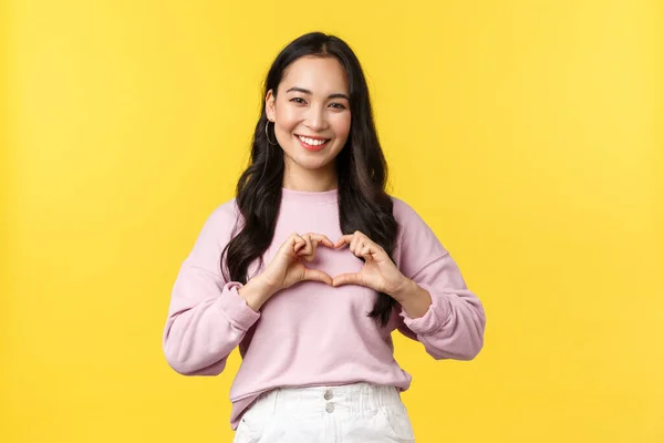 Estilo de vida, emoções e conceito de publicidade. Linda e carinhosa, menina coreana de aparência amigável sorrindo amplamente, mostrando sinal de coração sobre o peito e de pé fundo amarelo — Fotografia de Stock