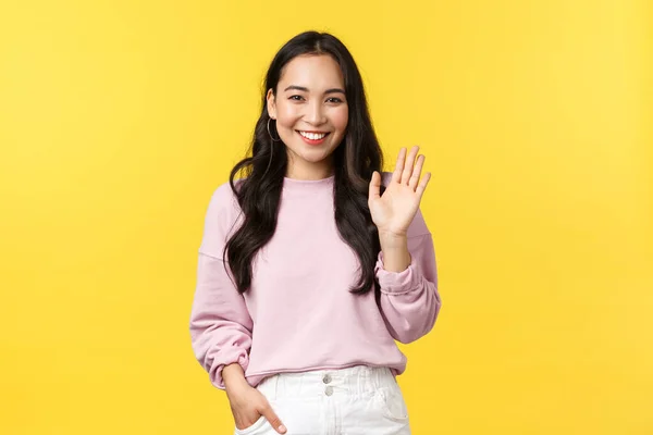 Lifestyle, Emotionen und Werbekonzept. Nette stilvolle asiatische Freundin winkt mit der Hand, um hallo zu sagen, lächelt freundlich zur Begrüßung, macht Hallo oder Willkommensgeste, gelber Hintergrund — Stockfoto