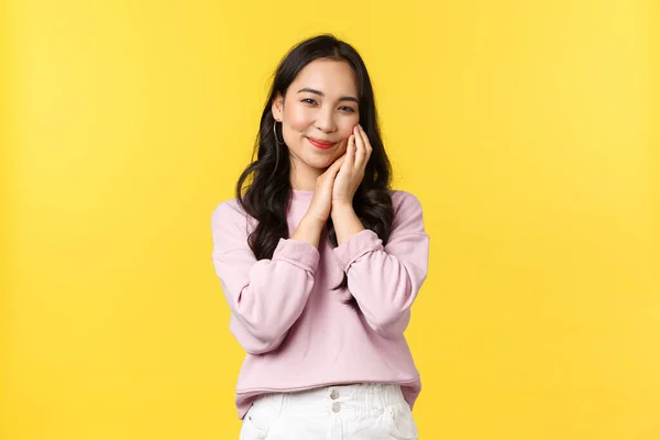 Estilo de vida, emoções e conceito de publicidade. Adorável sorrindo asiático mulher olhando tímido e bonito, tocando seu rosto com maquiagem em, sentindo-se bonito, de pé fundo amarelo — Fotografia de Stock