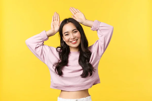 Estilo de vida, emoções e conceito de publicidade. bonito e bobo ásia menina fazendo coelho orelhas de mãos e dança, olhando kawaii com feixe branco sorriso, sinta feliz de pé amarelo fundo — Fotografia de Stock