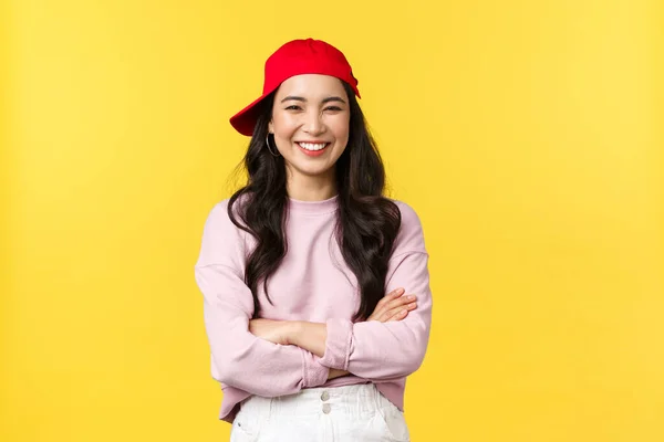 Az emberek érzelmei, életmód szabadidő és szépség koncepció. Boldog vidám ázsiai nő mosolyog és nevet, keresztbe karok mellkas, néz hűvös hipster piros sapka sárga háttér — Stock Fotó