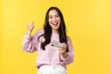 İnsanların duyguları, yaşam tarzı, eğlence ve güzellik konsepti. Hevesli Asyalı kız oyuncu cep telefonuyla oynuyor, akıllı telefonu oyun seviyesini geçmek için eğiyor, gülümsüyor, yumruk tokuşturarak kazanıyor, evet diyor.