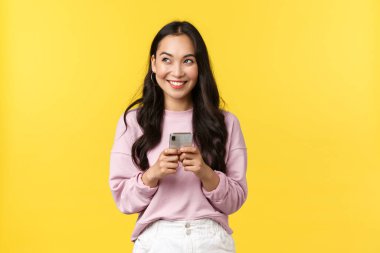 İnsanların duyguları, yaşam tarzı, eğlence ve güzellik konsepti. Hayalperest ve heyecanlı genç Asyalı kadın cep telefonu kullanıyor, gülümsüyor ve sol üst köşeye bakıyor.