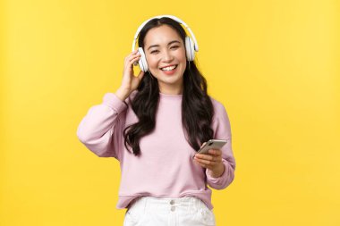 İnsanların duyguları, yaşam tarzı, eğlence ve güzellik konsepti. Mutlu gülümseyen Asyalı kadın kablosuz kulaklıkla müzik dinliyor ve güzel sesten memnun görünüyor, elinde cep telefonu var.