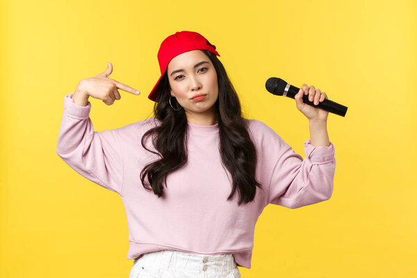 People emotions, lifestyle leisure and beauty concept. Cool and sassy stylish female raper, girl singer in red cap, acting confident, holding microphone and pointing at herself to show-off