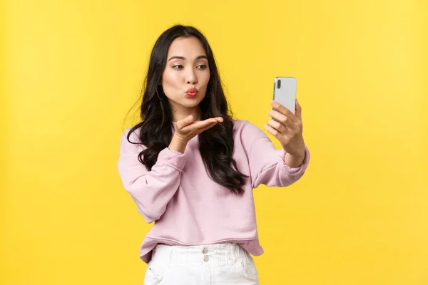 People emotions, lifestyle leisure and beauty concept. Stylish and flirty asian girl taking photo for social media, make selfie, sending air kiss at smartphone camera, stand yellow background — Stock Photo, Image