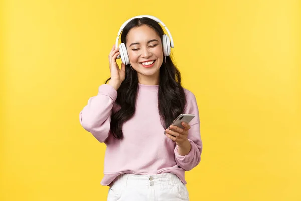 Emocje ludzi, wypoczynek w stylu życia i koncepcja piękna. Uśmiechnięta, nowoczesna azjatycka dziewczyna słuchająca muzyki w słuchawkach bezprzewodowych. Kobieta korzystających z ulubionej piosenki w słuchawkach, trzymając telefon komórkowy — Zdjęcie stockowe