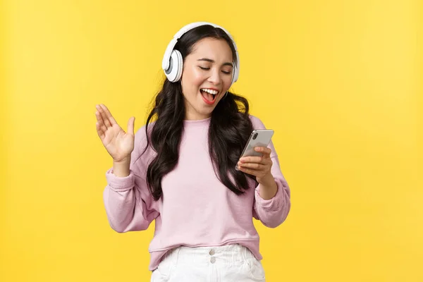 Gente emociones, estilo de vida ocio y concepto de belleza. Sonriendo feliz y despreocupado chica asiática cantando aplicación de karaoke en los auriculares, la lectura de la canción desde el teléfono móvil, de pie fondo amarillo — Foto de Stock