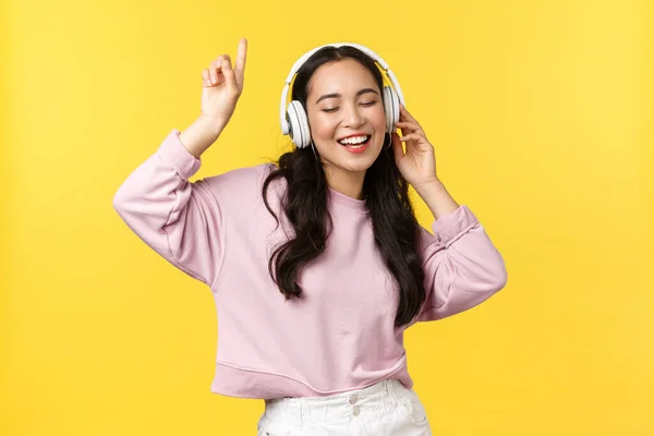 Gente emociones, estilo de vida ocio y concepto de belleza. Mujer asiática bastante feliz en auriculares inalámbricos, escuchando música y bailando, cantando, disfrutando del verano, de pie fondo amarillo — Foto de Stock