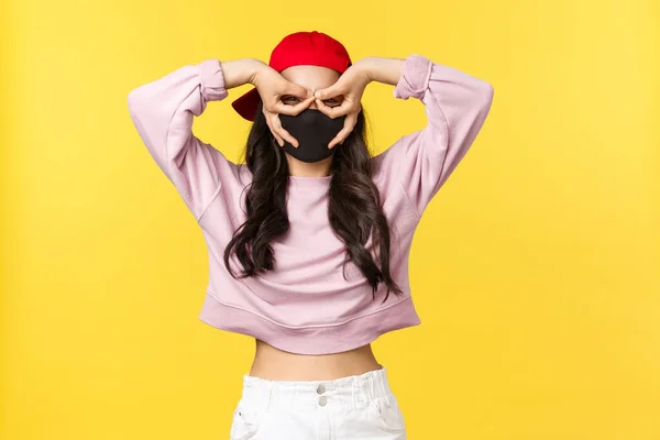 Covid-19, estilo de vida social-distanciamiento, prevenir el concepto de propagación del virus. Funny y linda chica asiática en máscara facial y gorra roja, hacer gafas falsas con los dedos sobre los ojos, mirada sorprendida e impresionada —  Fotos de Stock